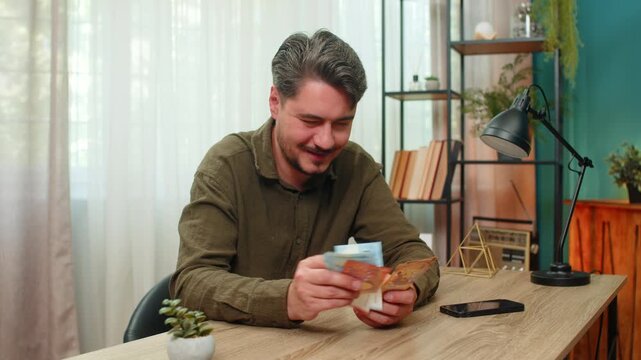 Middle-aged man at home counts cash smiling pleased by financial success feeling secure and proud. Young adult guy at table flips euro bills stacking money appreciating wealth gains after hard work