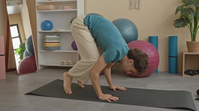 Young hispanic man holds crow pose on yoga mat using hands for balance in a studio surrounded by exercise balls and foam rollers; calm focus.