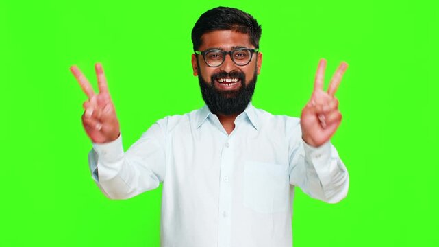 Indian man forms V sign with fingers friendly smile, expressing peace or triumph in confident open gesture. Arabian guy isolated on green chroma key background showing victory, success or harmony sign