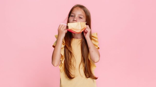 Little child girl fearfully shows a stop gesture while wearing a medical mask, then removes it and smiles with visible relief. School kid on pink background, portraying transition from fear to joy.