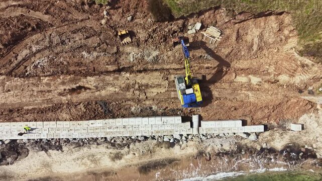 Heavy machinery works on land near the shore, placing large blocks in a line on the beach. The scene shows a mixture of dirt and ocean waves in the daytime.