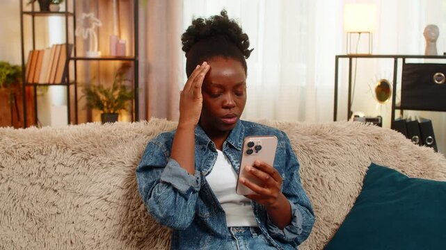 African American young woman looks disappointed while reading poor test results, losing an online game on smartphone. Black girl sighs deeply, sitting alone and overwhelmed by negative digital news.