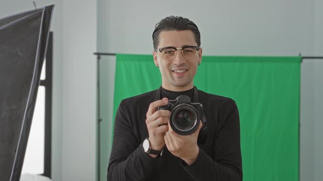 Young man photographer holding camera with both hands in a studio with green screen and softbox lighting, smiling at lens; creative confidence.
