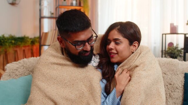 Indian couple man and woman sitting on home sofa, sharing soft hug under warm blanket in silence and comfort. Young family enjoying peaceful cozy moment together, feeling safe and emotionally close.