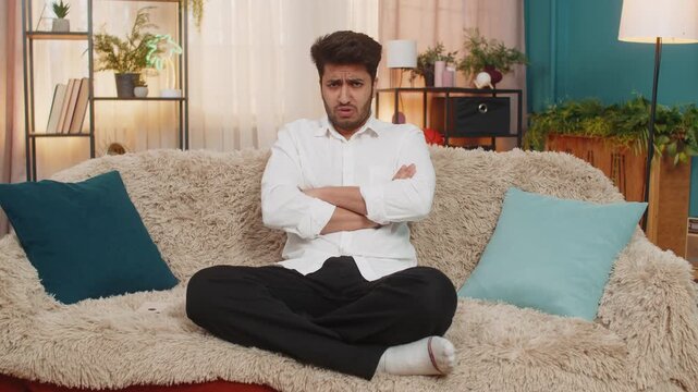 Indian man at home on sofa gripping smartphone tightly, upset, angrily tossing it aside after bad news. Arabian guy frustrated, annoyed, reacting emotionally, showing stress, disappointment irritation