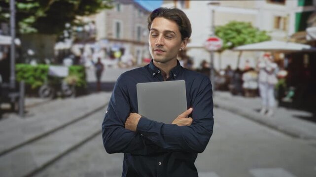 Man holding laptop with crossed arms on a street cafe with cobbles and tram tracks, looking sideways; contemplation.