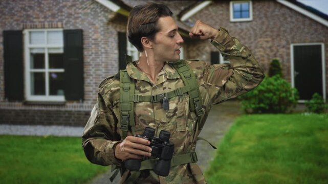 Man flexes arm and holds binoculars in front of building with brick facade, window shutters, grass lawn and stone path while wearing camouflage uniform and dogtags; pride duty.