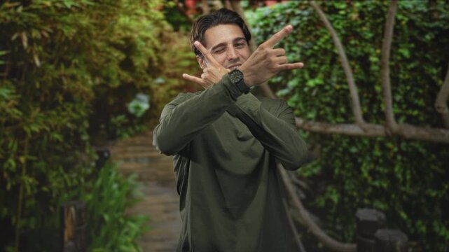 Man crossing arms and winking while making hand signs with watch and earring visible on a forest walkway; confidence.