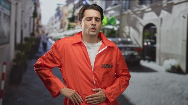 Man in orange jumpsuit clutching his stomach with hands over zipper and inmate number visible on a street lined with bollards and plants; suffering pain.