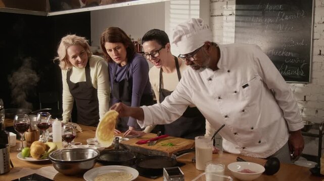 Tilt down shot of biracial chef putting oil to pan while teaching group of three women how to cook crepes during cooking master class in restaurant