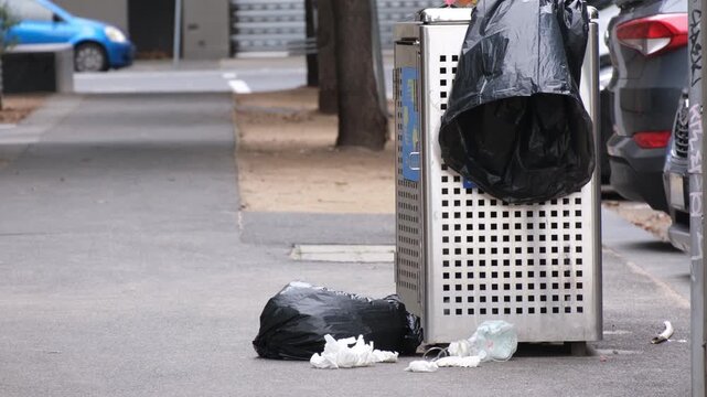 Overflowing stainless steel public rubbish bin on a Melbourne city street with garbage bags spilling onto the footpath pavement. Urban waste management issues and littering in Australia