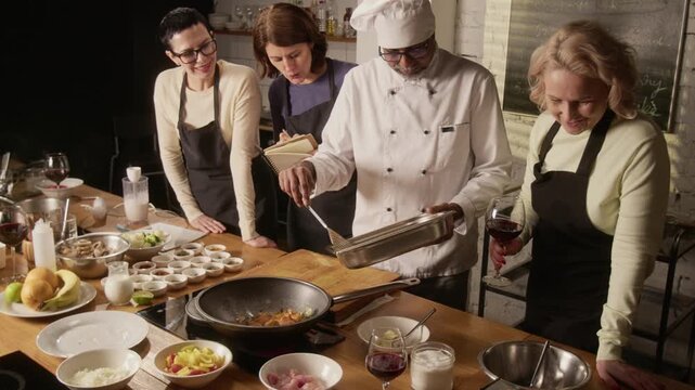 High angle shot of chef putting seasoned chicken into frying pan while group of women taking notes during cooking master class