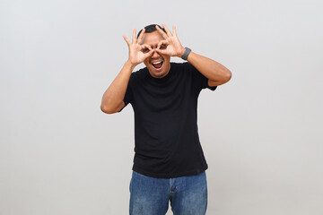Fototapeta premium Cheerful and happy adult man puts finger to eye, makes gesture of glasses or binoculars, looks at something in front, concept of advertising and offers, stands isolated on white background.