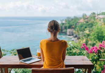 Fototapeta premium Woman sitting at a wooden table with a laptop and drink, enjoying remote work and freedom with a stunning ocean and green cliff view