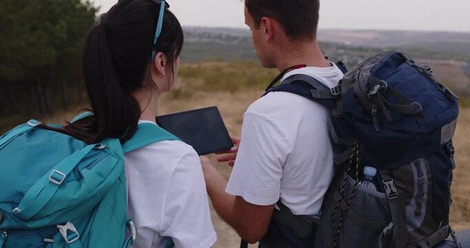 Hikers couple check tablet map for GPS navigation. Tourists stand on trail with backpack gear, planning route and checking signal to avoid getting lost in nature. Adventure and hiking concept. 