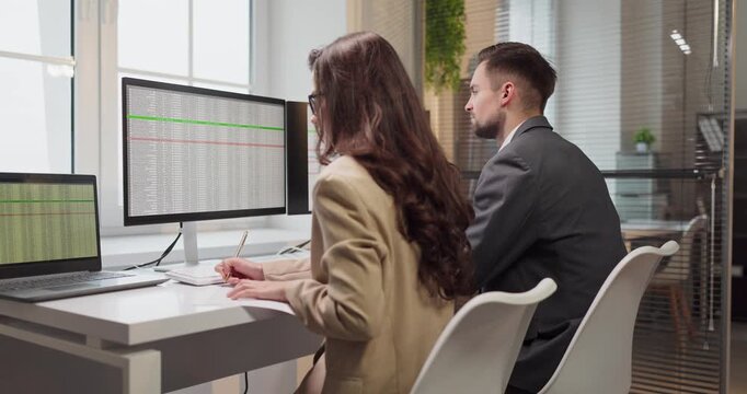 Analysts teamwork data on computer screens. Two accountants in a modern office monitoring finance spreadsheets and consulting on results together. Concept: financial data analysis teamwork.