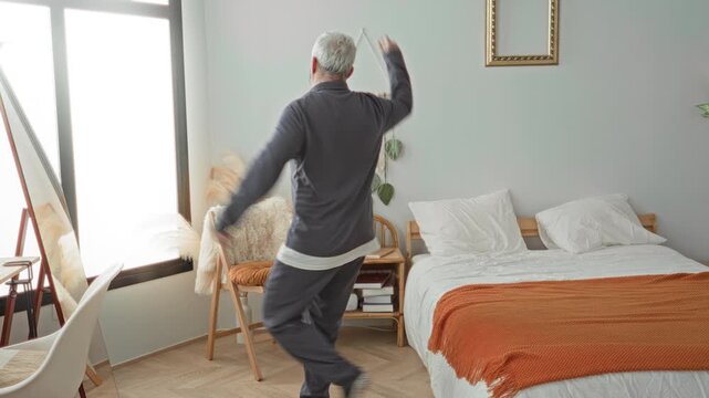 Middle age grey haired man dancing with arms outstretched beside an unmade bed with orange throw in a sunlit building; contentment morning.