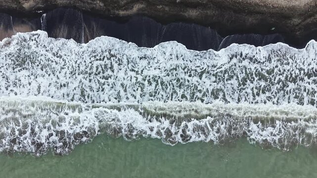 Waves roll onto a sandy beach as the tide comes in. The water is dark near the shore and lighter farther out, showing the movement of the sea. Clouds are present in the sky.