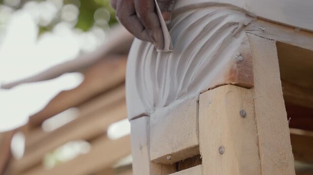 Closeup black hands shaping decorative corbel outdoors, commission woodworker performing carpentry finishing, steady sanding motion against timber support, sunlit bokeh, client project, tactile