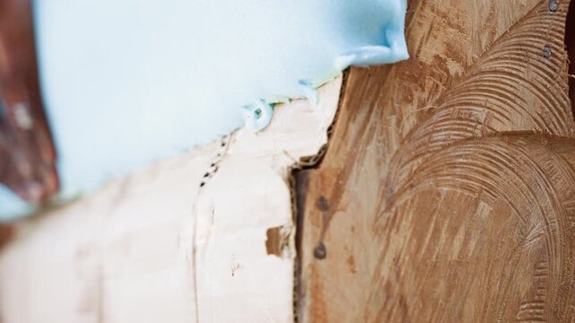 Black craftsman tacking foam to wooden frame, steady darkskinned hands pressing and aligning foam, nails and staples visible along seam, smoothing edge for snug fit, close handheld upholstery work