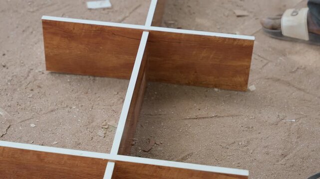 Wooden cross layout on sand showing two intersecting melamineedged boards on dusty ground, closeup of particleboard divider with subtle sawdust, precise joinery and measured placement for shelf