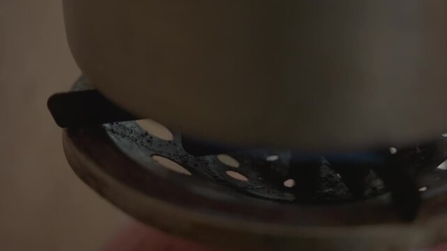 Pot placed over blue flame on burner, metal saucepan settles on rusted grate, flame intensifies beneath base, heat transfer begins, steam hint, practical kitchen ritual, handheld motion captured
