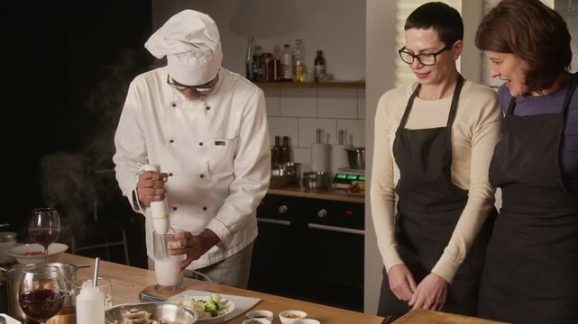 Biracial male chef using mixer while group of women paying attention while attending cooking master class in restaurant