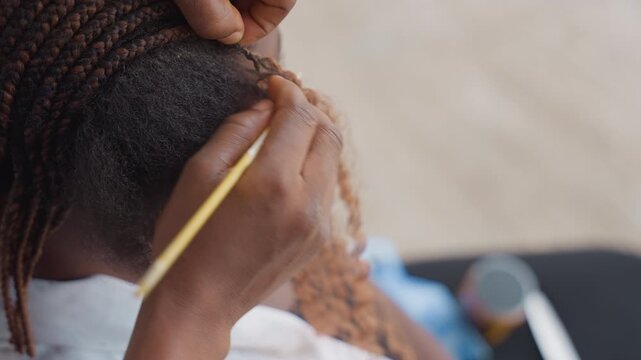 Closeup black stylist installing extensions using yellow hook, hands looping curly synthetic hair into cornrow base, controlled tension, focused technique, blurred workspace background, professional