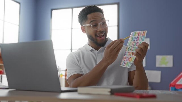 Man teacher pointing pencil at colorful alphabet sticker sheet while teaching via laptop in a building classroom; encouragement learning.