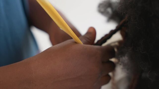 Closeup hands twisting black hair around yellow comb, careful sectioning and firm grip show meticulous technique midlength curls revealed, focused motion, concentrated rhythm, salon study atmosphere