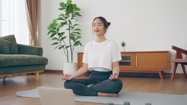 Young Asian woman sits cross-legged on the floor of her cozy home, watches an online yoga class on her laptop and meditates in yoga poses. She looks calm and relaxed in her sportswear