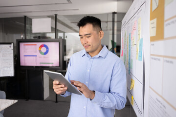 Fototapeta premium Project coordinator review. Confident young businessman stand near Kanban workflow model with tasks schedule on sticky notes check team work progress on digital tablet set deadlines priorities online