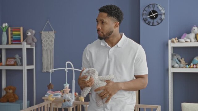 Man holding a gray plush toy over a wooden crib in a nursery, looking down while standing near shelves, clock and mobile; tenderness calm care.