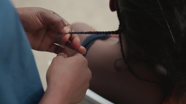 Closeup hands finishing braided end. Apprentice threading small bead onto braid near shoulder, fingers pinching knot, focused motion, sunlit skin, casual chair, patient energy, delicate finishing