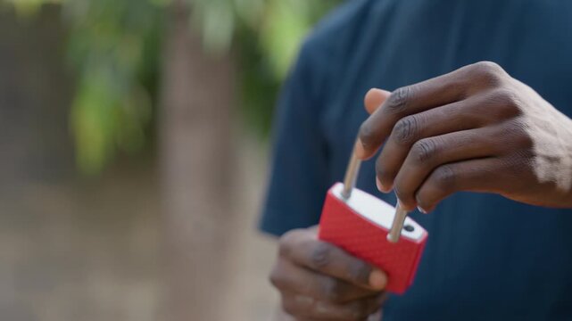 Closeup hands unlocking red padlock outdoors, slow deliberate motions reveal keys turning in metal shackle, natural light, blurred foliage background, tactile focus on fingers and texture, calm
