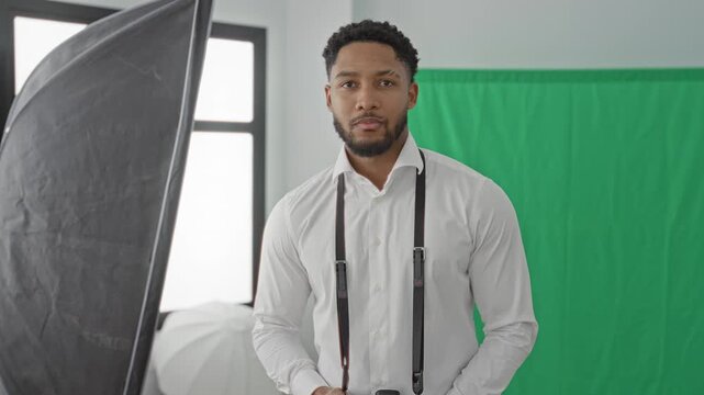 Man holding camera and smiling while aiming lens in studio with green screen and softbox lighting; creative confident.
