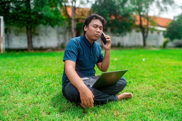 Obraz premium A person sitting on grass in a park, using a laptop and talking on a phone. The outdoor setting features greenery and trees, showcasing a remote work or study environment