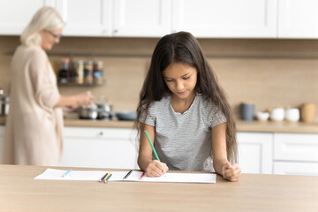 Focused little girl drawing pictures with colorful pencils in paper album while her older granny doing household chores, multigenerational people family spend weekend at home. Hobby, pastime activity
