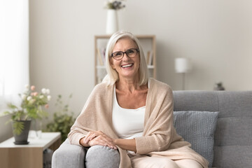In cozy living room mature female wear casual clothes posing seated on sofa, smile, looking at camera with joyful expression, enjoy carefree, untroubled life on retirement and pleasant rest at home