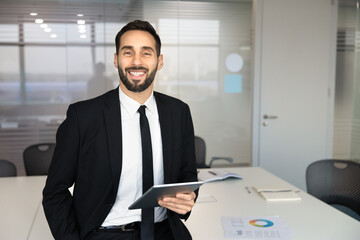 Office portrait of smiling businessman holding digital tablet, looking at camera, standing in boardroom. Represents professional workflow, effective corporate tasks management and business success