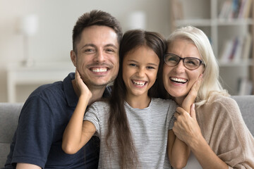 Pretty little 7s girl posing for photo with lovely older 60s grandma and young 35s handsome daddy, sitting on sofa looking at camera, laughing, feel happy. Multi-generational family people, portrait