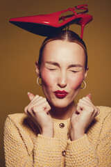 Young woman with closed eyes and red lipstick balances red high heel shoe on her head. She wears yellow sweater and gold hoop earrings on brown background. Concept of fashion and fun.