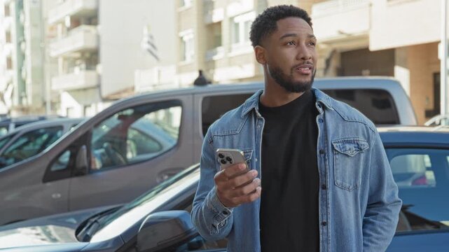 Man holding smartphone in right hand while glancing sideways near parked cars by apartment buildings on a street; thoughtful urban pause.