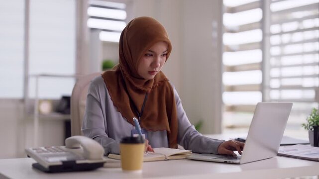 Focused Asian Muslim adult woman diligently working laptop writing notes professional modern office desk workspace productivity