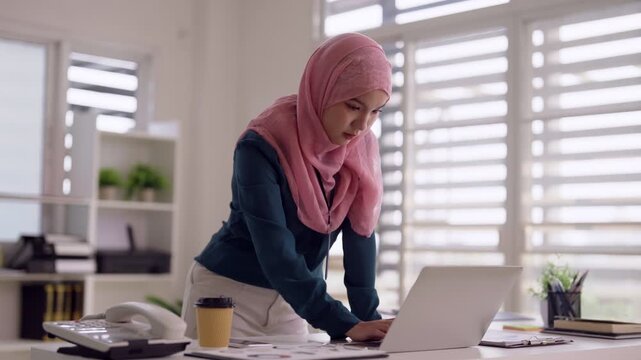 Adult Asian Muslim woman using laptop computer typing business reports working modern office focused remote work environment