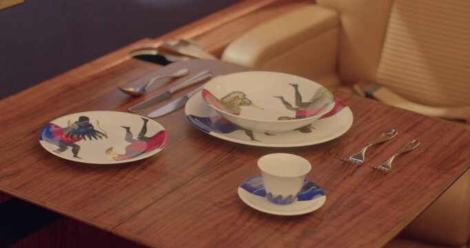 Aircraft tray table with porcelain plates and saucer, cutlery aligned on wooden tabletop beside leather seat, illustrated plate patterns and colorful silhouettes, warm ambient