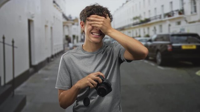 Boy covers eyes with hand while holding dslr camera, smiling with lens cap dangling, wearing gray t shirt on residential street lined with white townhouses and parked cars; shyness playfulness.