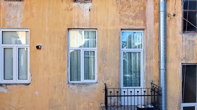Weathered yellowish brown facade of an old building with three windows, small balcony, and vertical pipe, showing peeling paint, discoloration, and signs of age