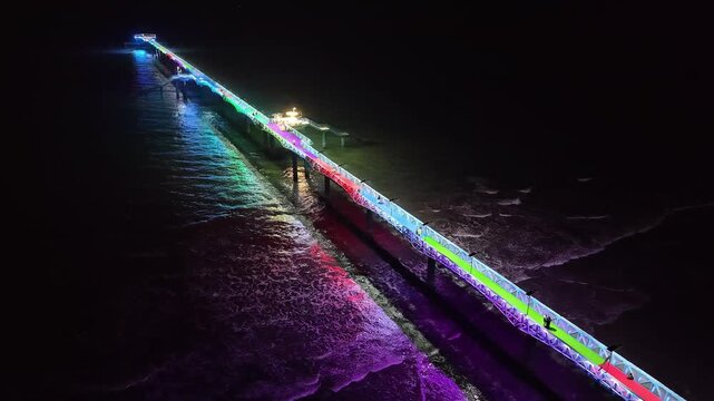 Night pier scene with colorful illuminated lights reflecting on water with waves at beach.