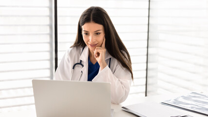 Focused female doctor sitting at desk in clinic office, reviewing medical information using laptop, consulting patient remotely, analyzing data, communicating online during telemedicine appointment
